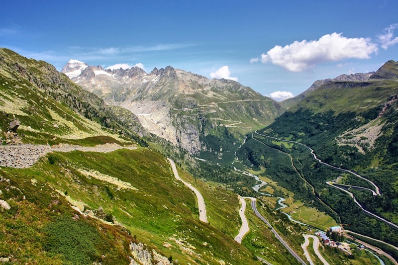 Kurvenreiche Passstrasse zwischen grünen Alpenhängen, dahinter Gletscher und Gipfel